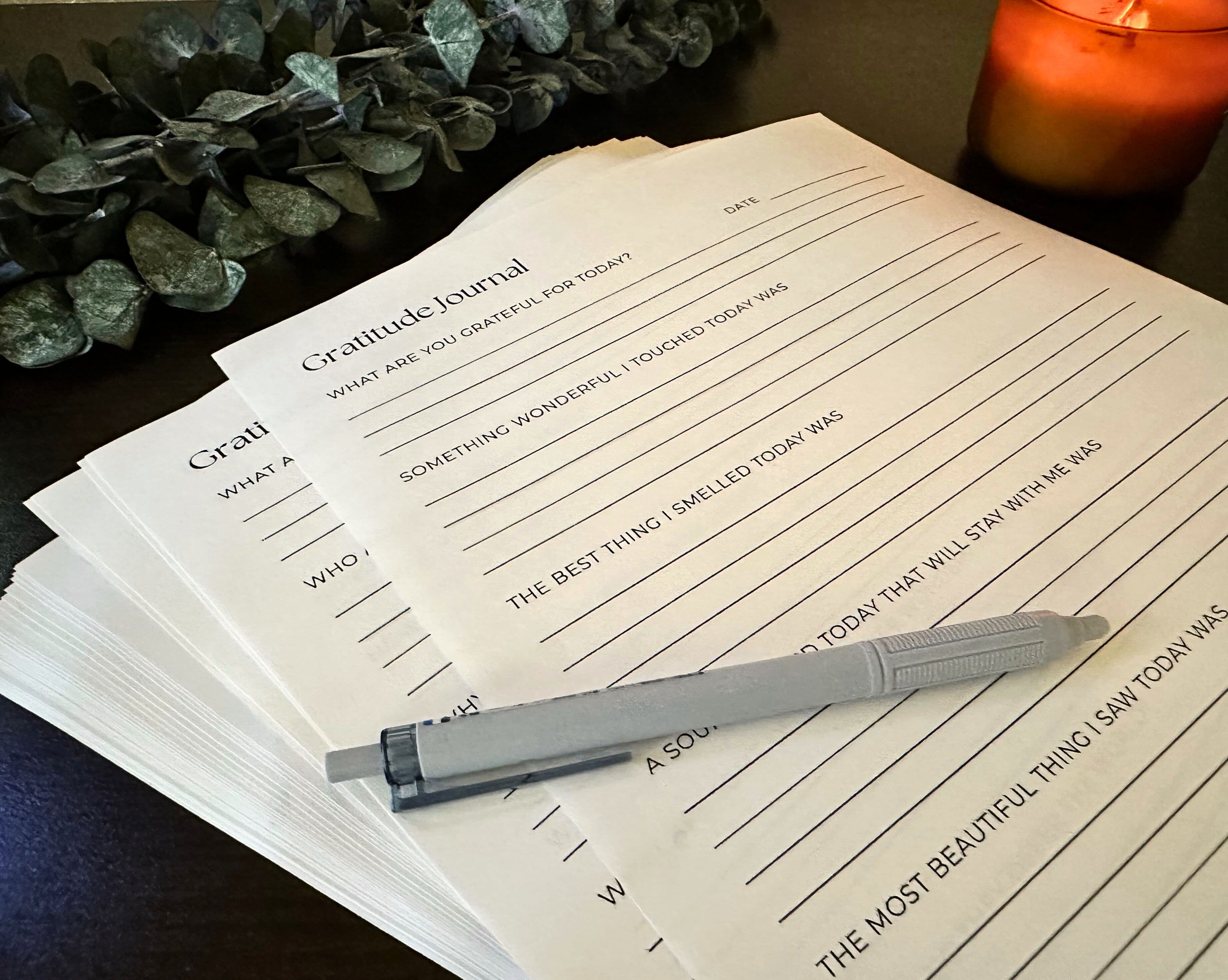 A stack of printable gratitude journal pages with a pen and candle, styled on a wooden desk for a cozy, reflective scene.