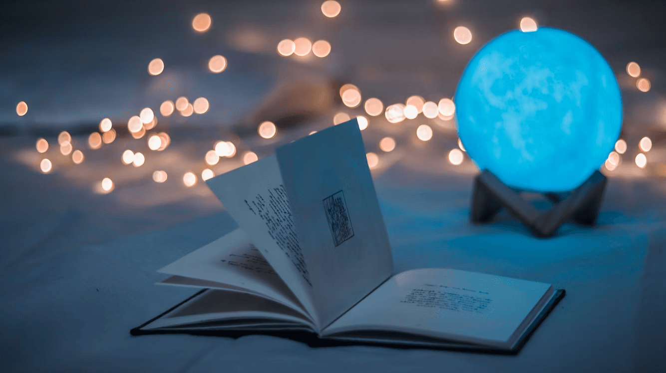Open book on a soft surface under moonlight, surrounded by glowing fairy lights and a blue orb lamp — a calm, mystical scene symbolizing ritual and reflection.